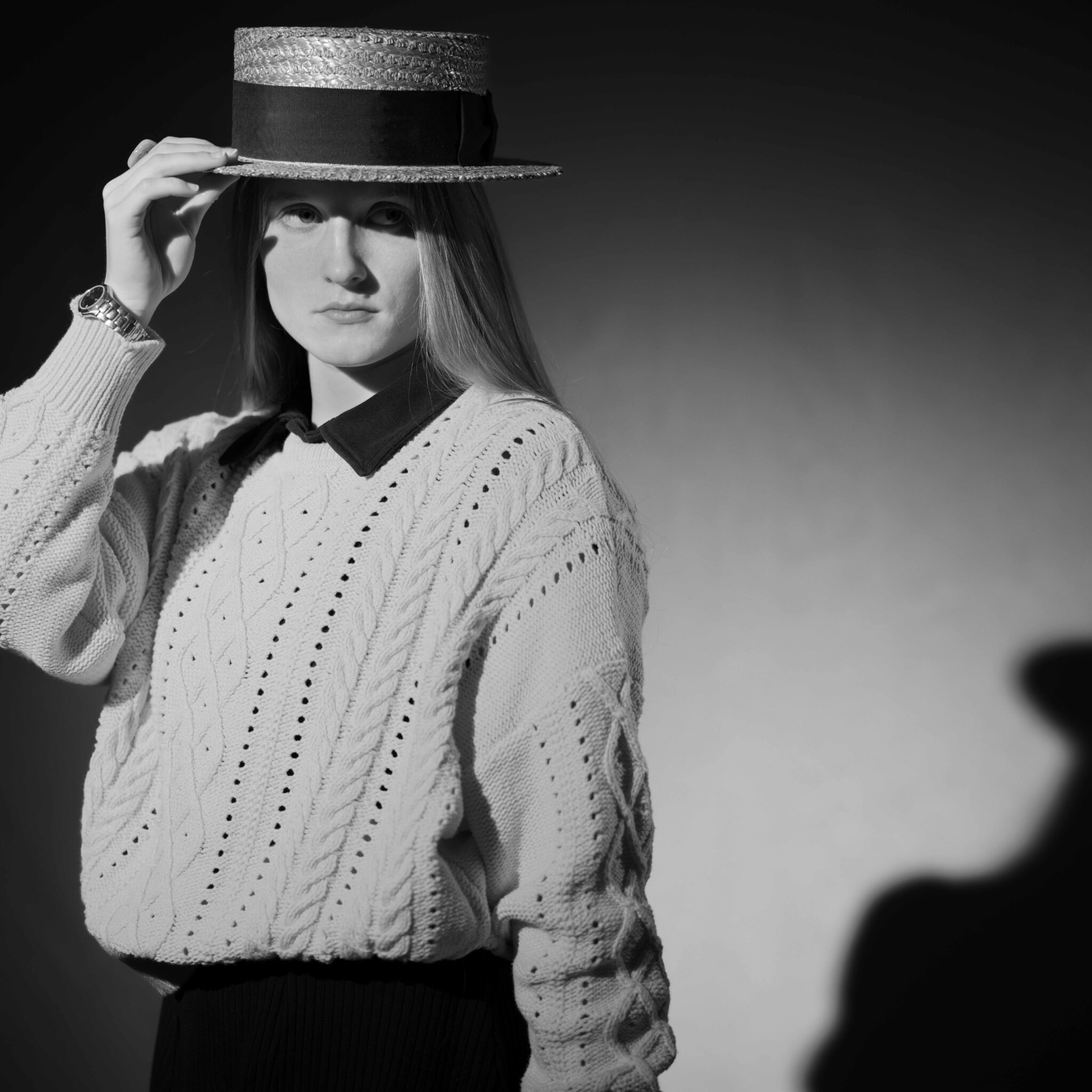 De Angelis Fotografica. Junge Dame mit einem Sonnenhut steht in hartem Lichtkegel und hält mit einer Hand ihren Hut. Sie ist elegant gekleidet.