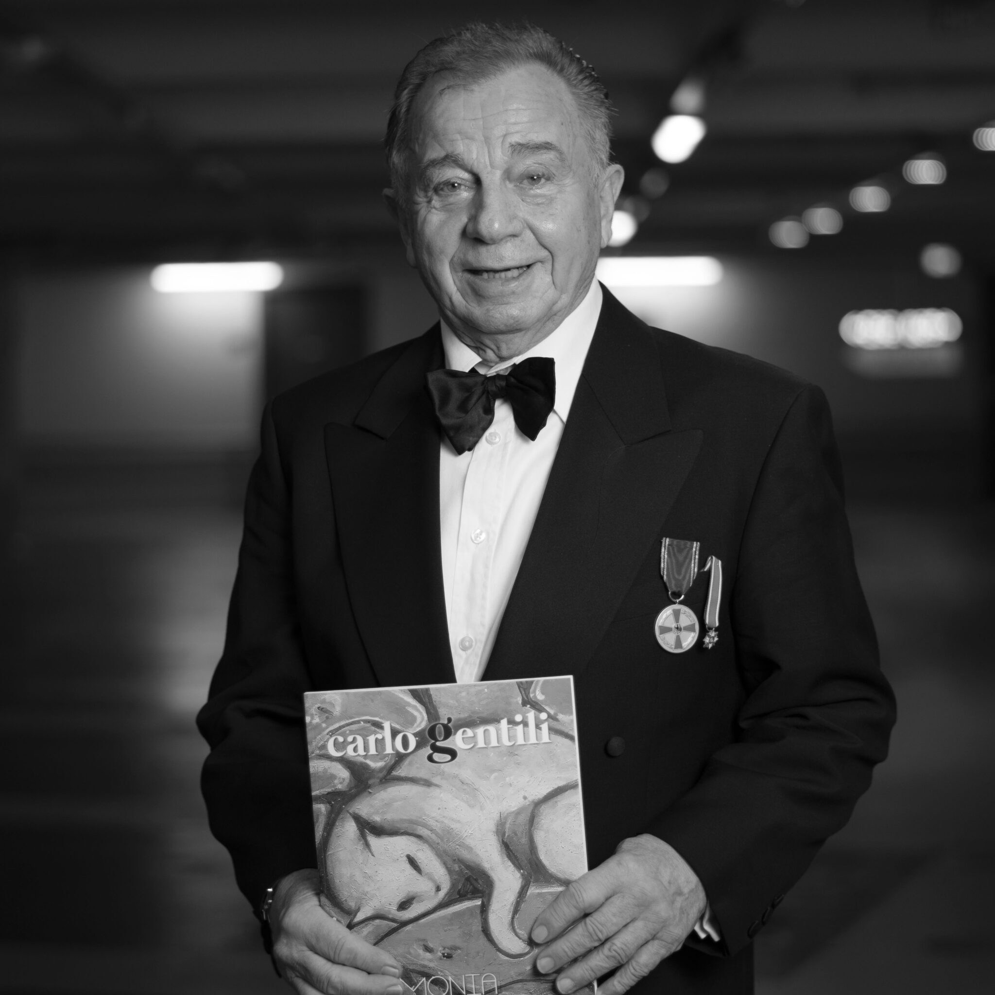 De Angelis Fotografica. Eleganter Gentleman mit Ehrung steht freundlichen lächelnd zur Kamera gewandt und hält ein Buch in der Hand.