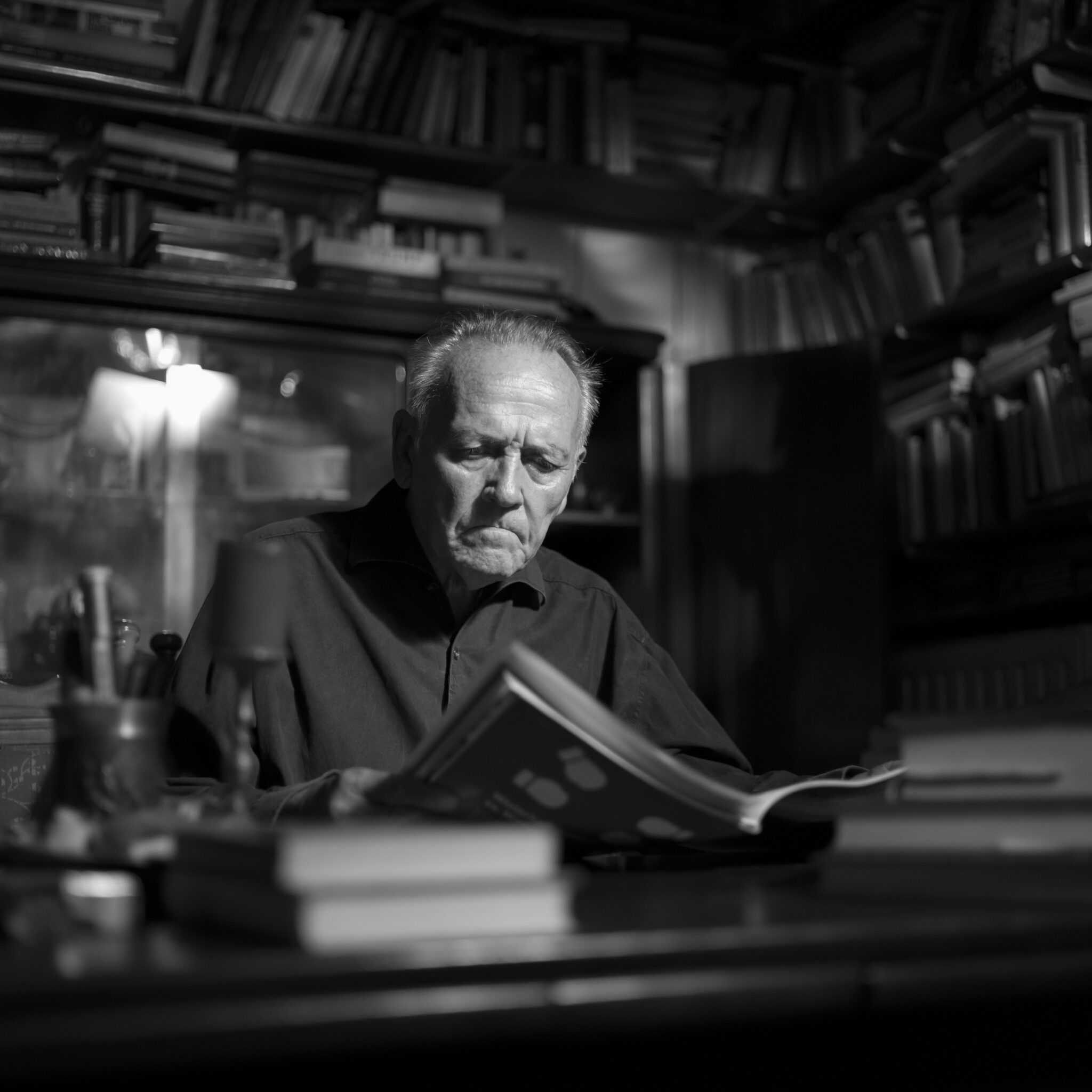 De Angelis Fotografica. Mann sitzt in einer Bibliothek voll mit Büchern an einem Tisch und schaut lesend auf ein Buch nach unten.
